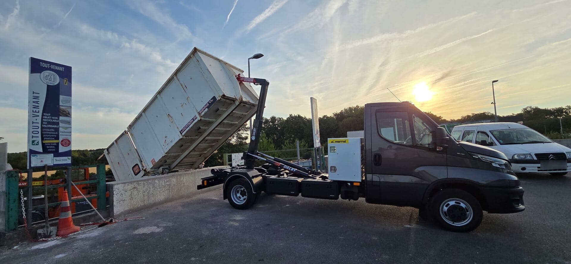 Camion à bras levant une benne blanche au-dessus du quai tout-venant d’une déchèterie, au coucher du soleil