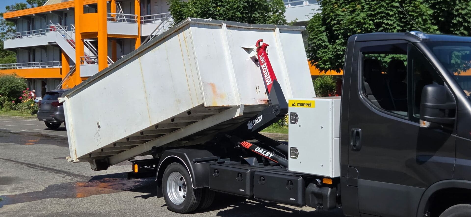Camion à bras basculant une benne blanche sur un parking, devant un immeuble aux balcons orange