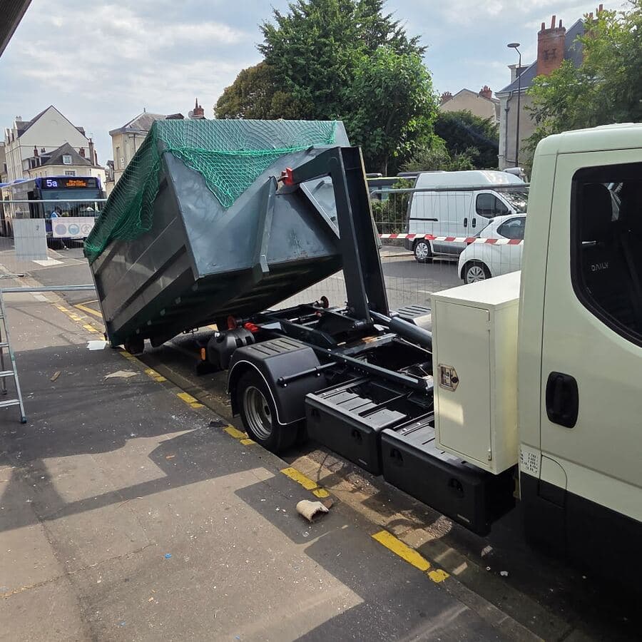 Camion à bras hissant une benne grise couverte d’un filet vert, dans une rue en ville, un bus à l’arrière-plan